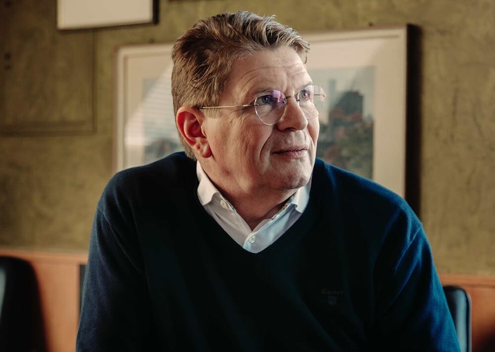 Ein älterer Mann mit Brille und kurzen, hellbraunen Haaren sitzt im Haus, trägt ein weißes Kragenhemd unter einem dunklen Pullover und schaut nachdenklich zur Seite, als träume er von der Freiheit, die er im Segelfliegen findet. Hinter ihm hängt ein gerahmtes Kunstwerk an der Wand.