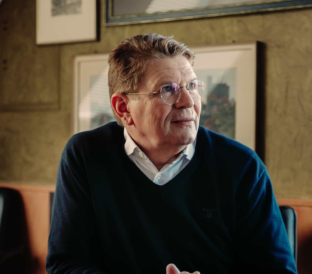 Ein älterer Mann mit Brille und kurzen, hellbraunen Haaren sitzt im Haus, trägt ein weißes Kragenhemd unter einem dunklen Pullover und schaut nachdenklich zur Seite, als träume er von der Freiheit, die er im Segelfliegen findet. Hinter ihm hängt ein gerahmtes Kunstwerk an der Wand.