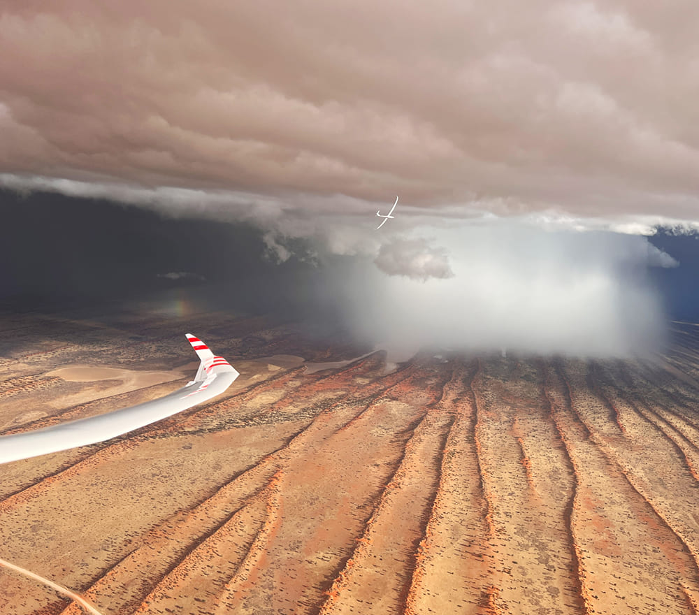 Blick aus einem Segelflugzeug während des Segelfliegens über eine trockene, zerklüftete Wüstenlandschaft unter Gewitterwolken und Regen, mit einem anderen, weit entfernten Segelflugzeug in der Nähe des Horizonts - ein Erlebnis von purer Freiheit.