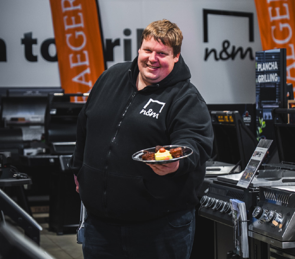 Ein lächelnder Mann in einem schwarzen Kapuzenpulli steht in einem Innenraum, hält einen Teller mit gegrilltem Essen in der Hand und bietet ihn an. Hinter ihm stehen Grills, orangefarbene Banner und das lebendige Ambiente des Grillen Genussquartier Niemeyer.