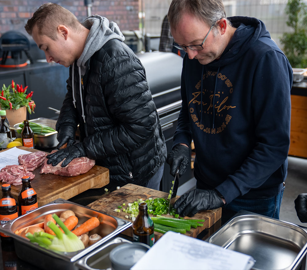 Zwei Männer in Handschuhen und Kapuzenjacken bereiten im Grillen Genussquartier Niemeyer Speisen im Freien zu. Einer schneidet grüne Zwiebeln und Sellerie, der andere rohes Fleisch. Auf dem Tisch daneben stehen frisches Gemüse und Bierflaschen.
