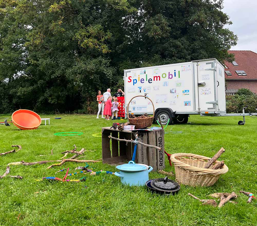 Eine grasbewachsene Wiese mit verstreutem Spielzeug, Eimern und Stöcken im Vordergund. Im Hintergrund ein weißes Spielemobil des Kinderschutzbundes Ammerland und mehrere Personen, die sich unter Bäumen versammeln und Familien in der Nähe eines Hauses Hilfe anbieten.