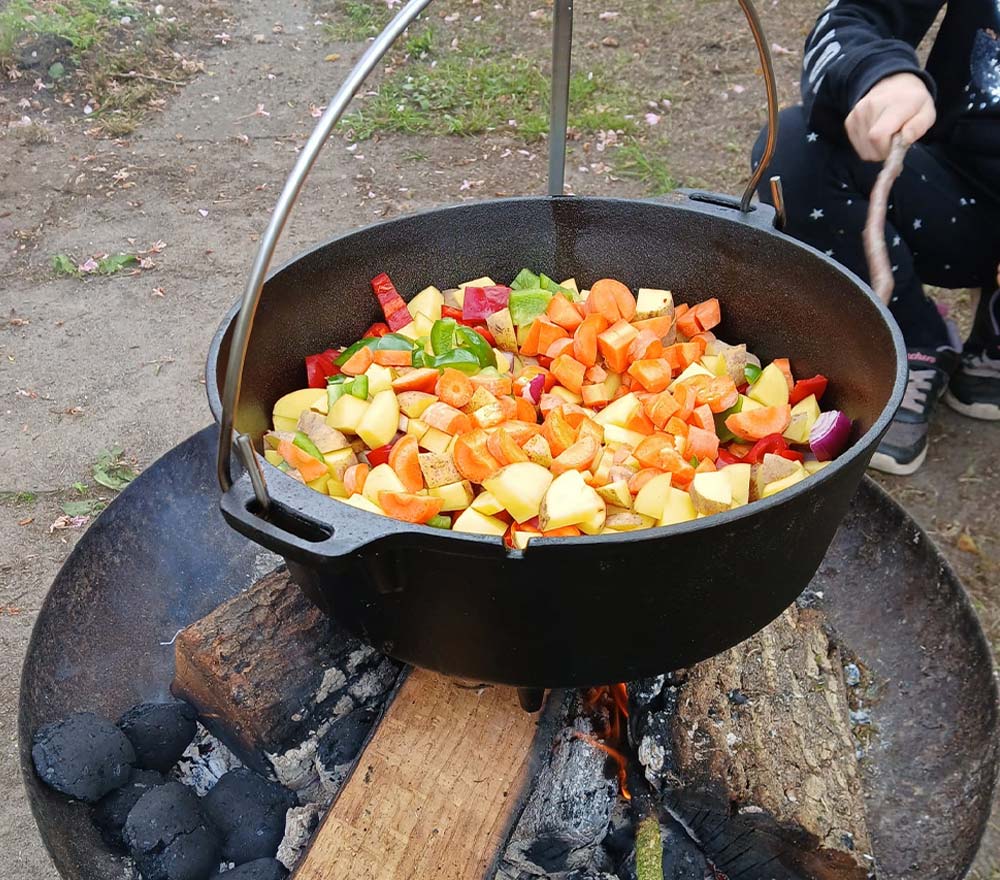 Ein schwarzer Topf mit kleingeschnittenem Gemüse wie Karotten, Kartoffeln und Paprika kocht über einem Feuer im Freien auf einem Grill. In der Nähe hilft ein Mitarbeiter des Kinderschutzbundes Ammerland den Familien dabei, das Erlebnis zu genießen, während Holz und Holzkohle die Flammen anheizen.