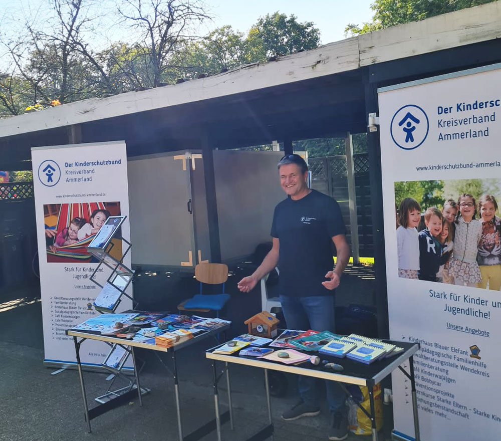 Ein Mann steht lächelnd hinter Tischen mit Broschüren und Artikeln an einem Außenstand des Kinderschutzbundes Ammerland. Auf Bannern sind Informationen und Fotos zu sehen, die für die Hilfe für Kinder und Familien der Organisation werben.
