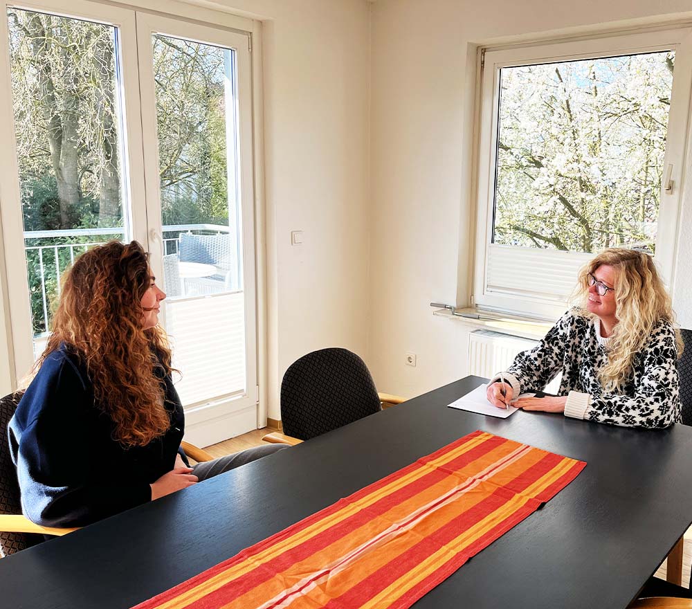 Zwei Frauen sitzen sich an einem dunklen Tisch mit einem bunt gestreiften Läufer gegenüber, in einem hellen Raum mit großen Fenstern und Bäumen draußen. Die eine macht sich Notizen, die andere hört zu - und bietet Hilfe für Familien beim Kinderschutzbund Ammerland an.