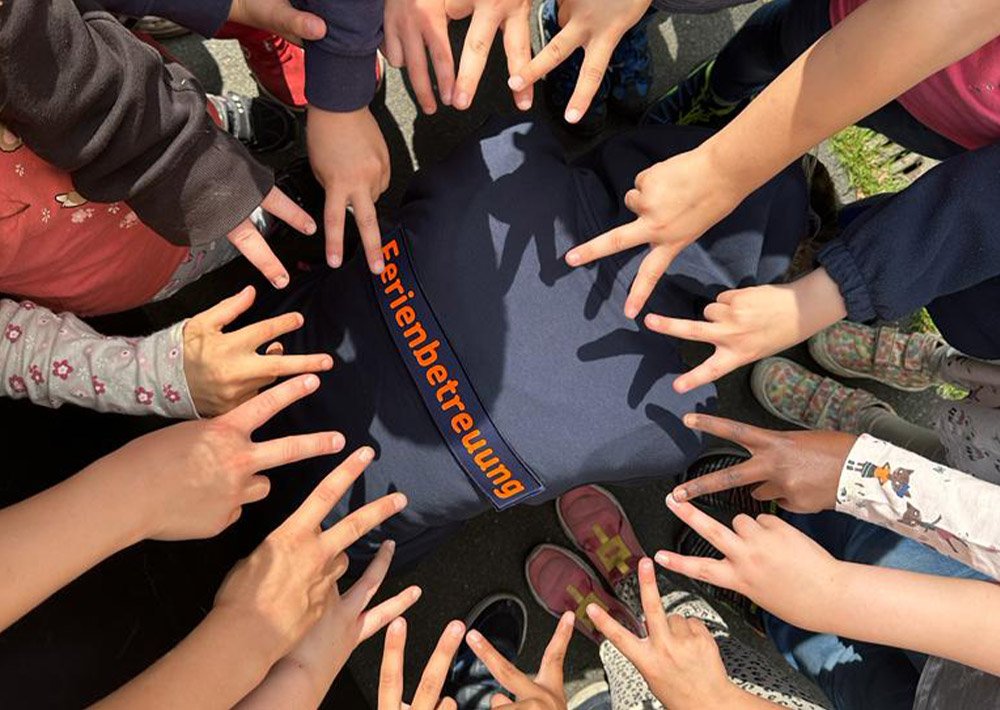 Eine Gruppe von Kindern zeigt mit den Fingern nach innen und formt Friedenszeichen um eine Person, die ein "Ferienbetreuung"-Shirt trägt, auf dem nur Hände, Arme und teilweise Beine zu sehen sind - ein Zeichen für die Unterstützung und Hilfe für Familien durch den Kinderschutzbund Ammerland während der Ferienbetreuung.