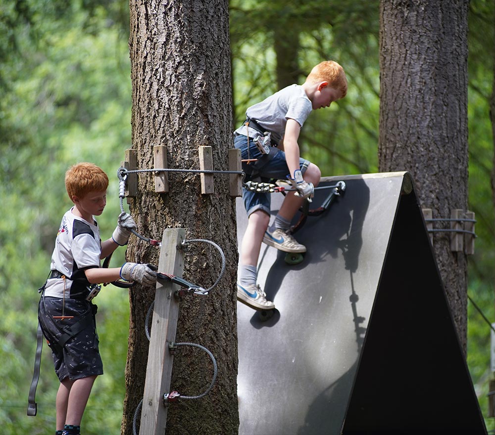 Zwei rothaarige Jungen mit Klettergurten und Handschuhen erleben im Kletterwald Thüle ein Seilabenteuer im Freien - der eine klettert eine Holzleiter hinauf, der andere erklimmt eine schräge Wand, was diesen Lieblingsort zum perfekten Waldspaß macht.