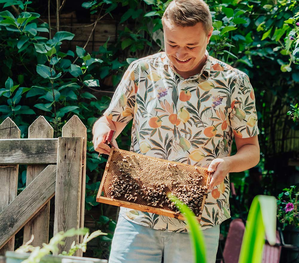 Ein lächelnder Imker in einem geblümten Hemd hält in einem üppigen Garten einen hölzernen Bienenstockrahmen, der mit Bienen bedeckt ist. Er steht in der Nähe eines Holzzauns und ist von grünen Pflanzen umgeben - und feiert die Kunst der Imkerei.
