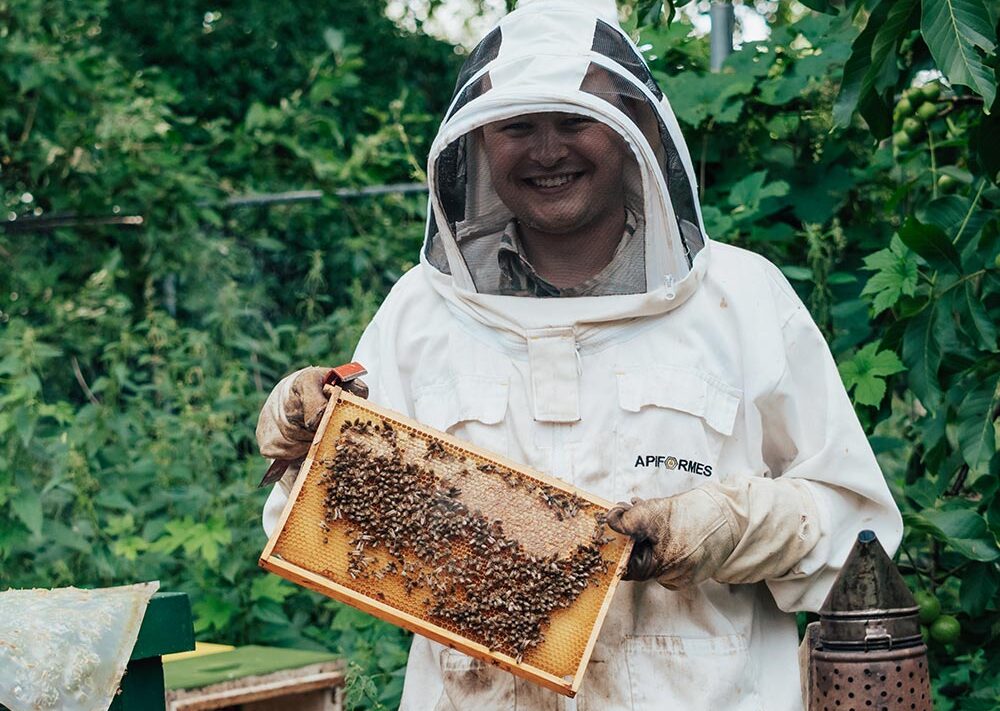 Ein lächelnder Imker im Schutzanzug hält einen mit Bienen bedeckten Wabenrahmen. Er steht im Freien neben der Bohmann-Imkereiausrüstung und ist von grünem Laub umgeben.