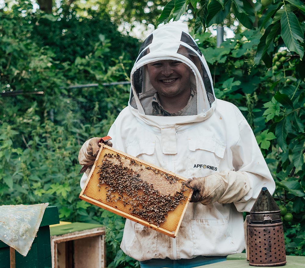 Ein lächelnder Imker im Schutzanzug hält einen mit Bienen bedeckten Wabenrahmen. Er steht im Freien neben der Bohmann-Imkereiausrüstung und ist von grünem Laub umgeben.