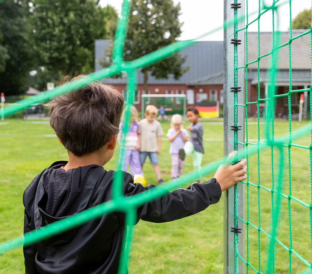 Ein Kind steht neben einem grünen Fußballtor und beobachtet eine Gruppe von Kindern, die gemeinsam auf einer Wiese im Oldenburger Land spielen. Hinter ihnen sind Bäume und Gebäude zu sehen, die die Wirkung der Regionalen Stiftung LzO Förderung in der Gemeinde verdeutlichen.