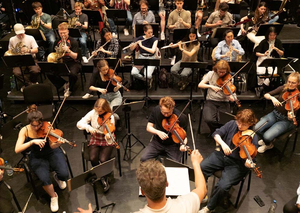 Ein Dirigent leitet ein großes Orchester mit Streich- und Blasinstrumenten während einer Probe auf der Bühne, von oben gesehen - eine Veranstaltung, die von der Regionalen Stiftung LzO Förderung Oldenburger Land unterstützt wurde.