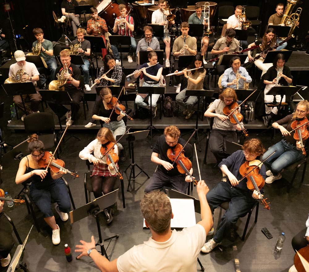 Ein Dirigent leitet ein großes Orchester mit Streich- und Blasinstrumenten während einer Probe auf der Bühne, von oben gesehen - eine Veranstaltung, die von der Regionalen Stiftung LzO Förderung Oldenburger Land unterstützt wurde.