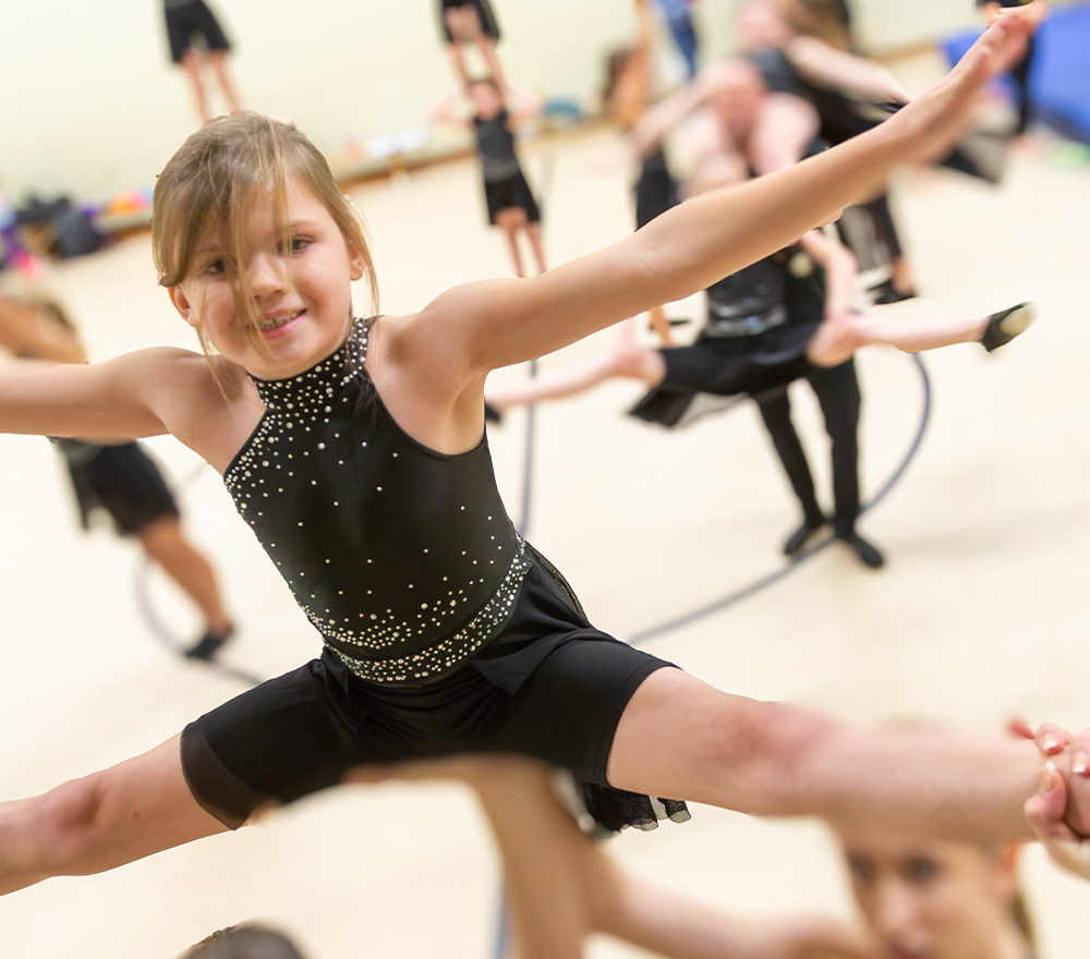 Ein junges Mädchen in einem schwarzen Kostüm mit Glitzern führt einen Spagatsprung während einer Gymnastik- oder Tanzübung in einer Halle aus, die von der Regionalen Stiftung LzO Förderung Oldenburger Land unterstützt wird, wobei andere Darsteller im Hintergrund verschwimmen.