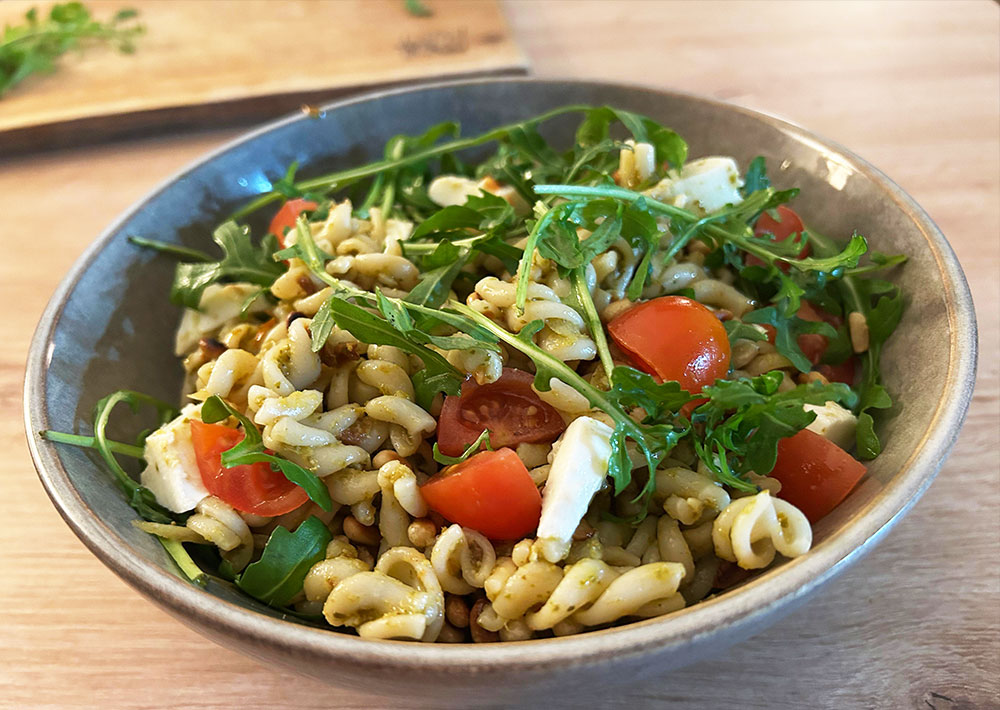 Eine Schüssel Nudelsalat mit Fusilli, halbierten Kirschtomaten, Rucolablättern und Mozzarella-Stückchen - perfekt für Sommerrezepte oder als Beilage beim Grillfest, serviert auf einem rustikalen Holztisch.