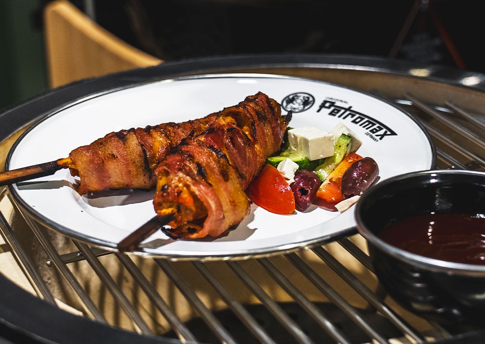 Zwei mit gegrilltem Speck umwickelte Spieße liegen auf einem weißen Teller mit einem frischen Sommersalat aus Tomate, Feta, Gurke und Oliven. Perfekt für Rezepte Grillen auf dem nächsten Grillfest, mit einer Schale dunkler Soße in der Nähe und dem Teller auf einem Metallgrill.