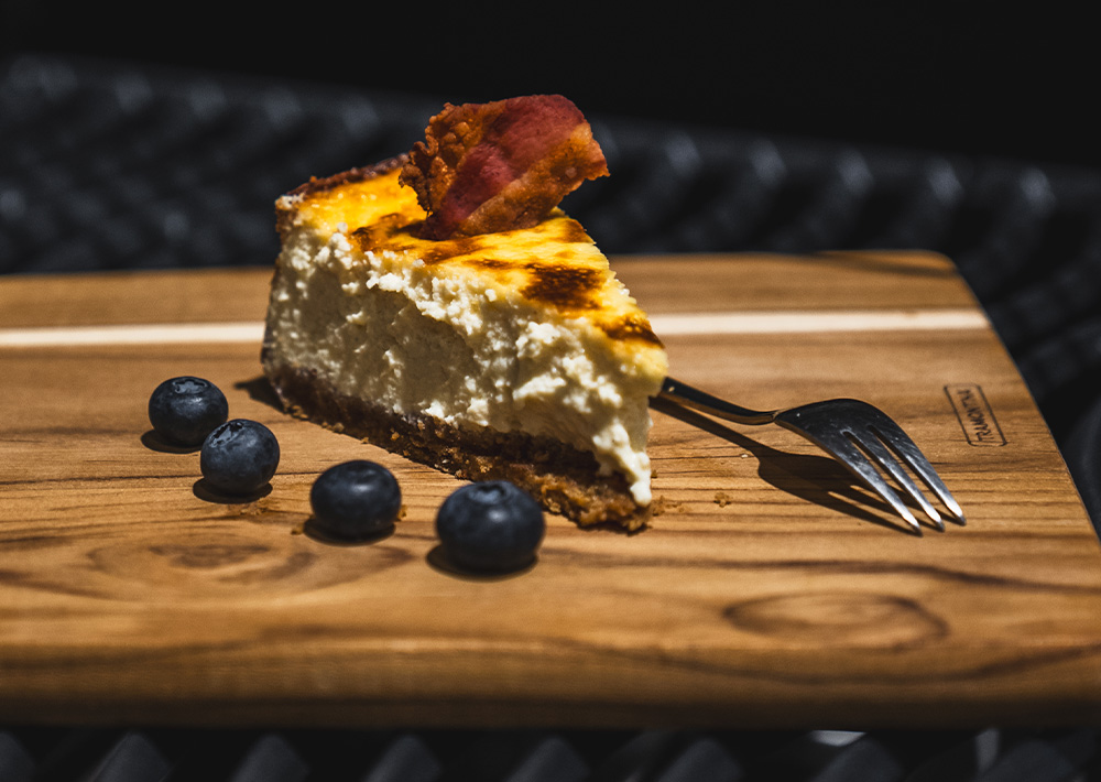Ein Stück Käsekuchen, belegt mit Speck, liegt auf einem Holzbrett, daneben eine Gabel und drei Blaubeeren. Dieses einzigartige Dessert passt perfekt zu den Sommer-Grill-Rezepten und ist ein echter Hingucker auf jedem Grillfest. Der Hintergrund ist dunkel und unscharf.