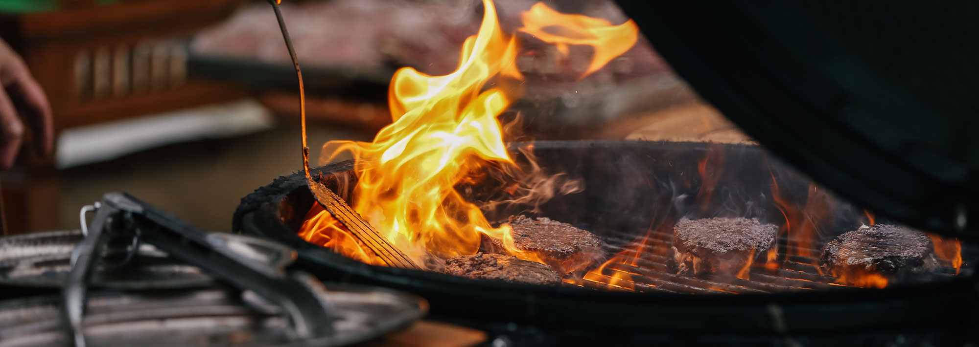 Fünf Hamburger-Patties werden auf einem Grill mit hellen Flammen gegart, perfekt für Sommer Grillfest Rezepte, während eine Hand ein Werkzeug oder einen Pinsel über das Feuer hält; im Vordergrund sind Grillwerkzeuge zu sehen.
