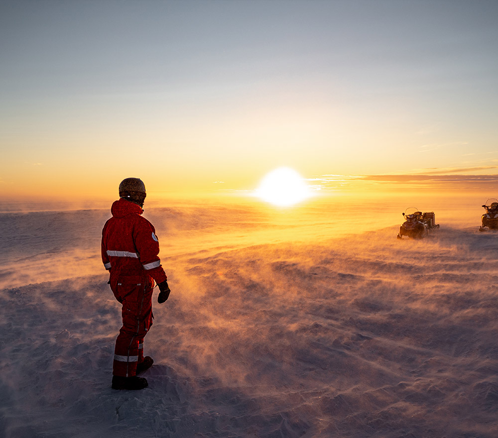 Eine Person in roter Winterkleidung steht in einer Schneelandschaft bei Sonnenuntergang, mit Schneeverwehungen in der Luft und Motorschlitten in der Ferne, unter einem goldenen Himmel - und fängt den Geist der Polarforschung Trautmann Antarktis ein.