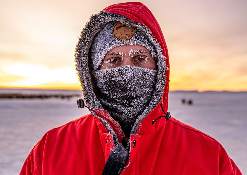 Eine Person in einem roten Parka mit pelzgefütterter Kapuze, Hut und Gesichtsmaske steht draußen in einer Schneelandschaft, die an die Polarforschung Trautmann Antarktis erinnert. Frost bedeckt ihre Augenbrauen, während im Hintergrund die Sonne untergeht.