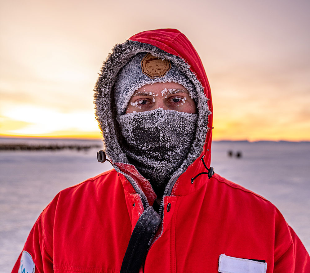 Eine Person in einem roten Parka mit pelzgefütterter Kapuze, Hut und Gesichtsmaske steht draußen in einer Schneelandschaft, die an die Polarforschung Trautmann Antarktis erinnert. Frost bedeckt ihre Augenbrauen, während im Hintergrund die Sonne untergeht.