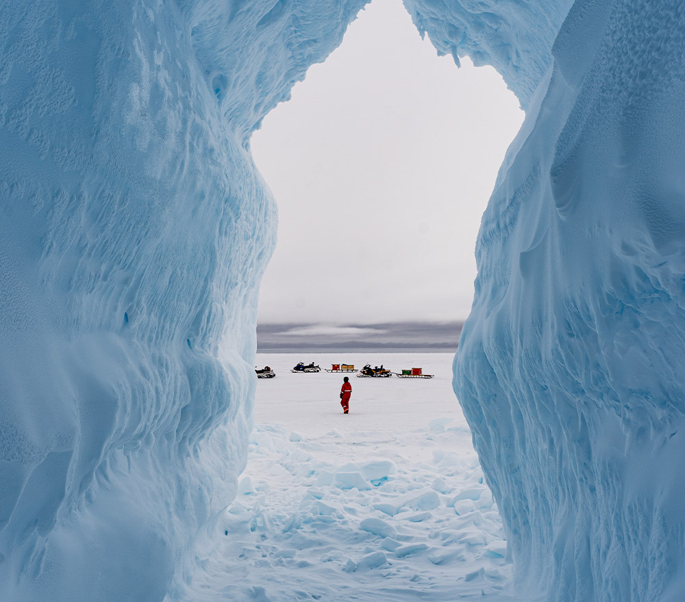 Eine Person in einem roten Mantel steht auf verschneitem Boden, umrahmt von hohen, gebogenen blauen Eiswänden - eine eindrucksvolle Szene der Polarforschung in der Antarktis. In der Ferne sind unter einem wolkenverhangenen Himmel Motorschlitten und Geräte zu sehen, die an Trautmanns Expeditionen erinnern.