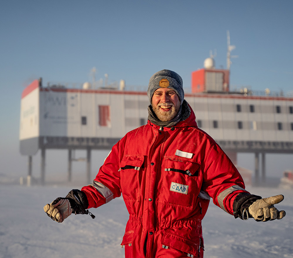 Eine Person in rotem Winteranzug und grauer Mütze steht mit weit ausgebreiteten Armen lächelnd in einer verschneiten Landschaft vor der hochgelegenen Forschungsstation Polarforschung Trautmann Antarktis.