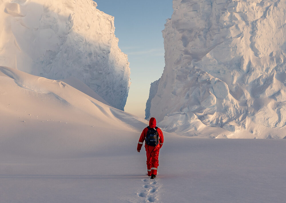 Eine Person im roten Schneeanzug und mit Rucksack begibt sich auf ein Abenteuer und wandert durch den Tiefschnee zu einer schmalen Passage zwischen hoch aufragenden Eisfelsen unter einem klaren Himmel.