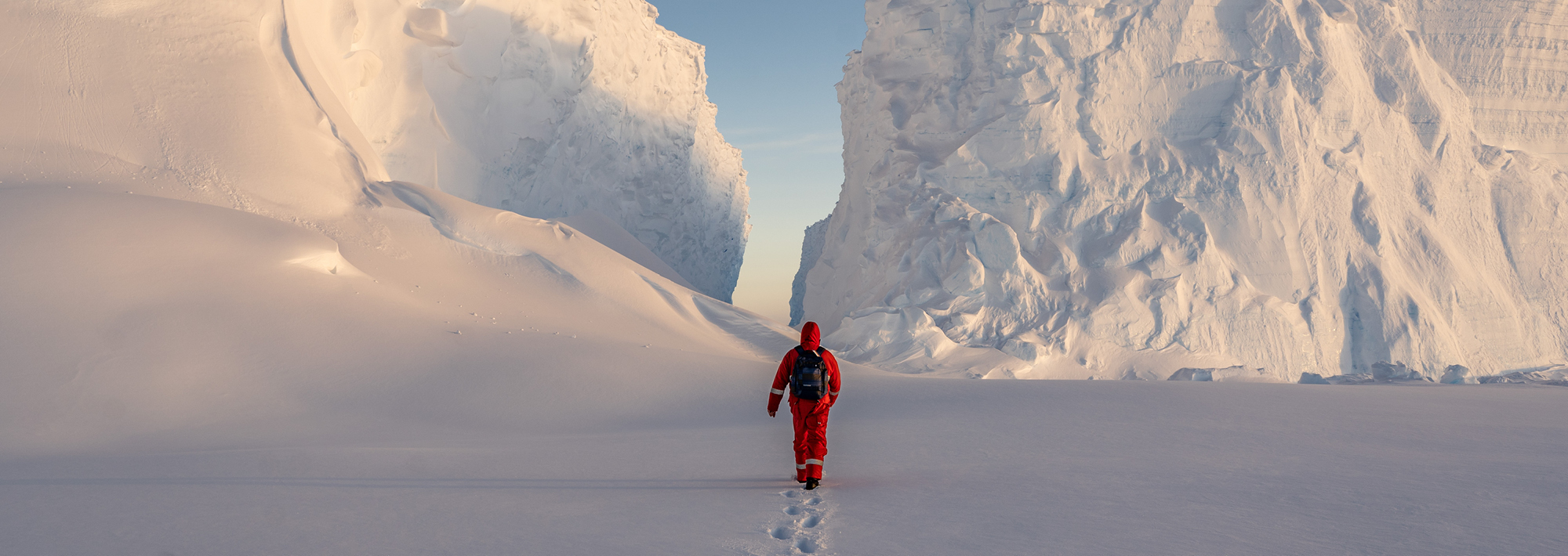 Eine Person im roten Schneeanzug und mit Rucksack begibt sich auf ein Abenteuer und wandert durch den Tiefschnee zu einer schmalen Passage zwischen hoch aufragenden Eisfelsen unter einem klaren Himmel.