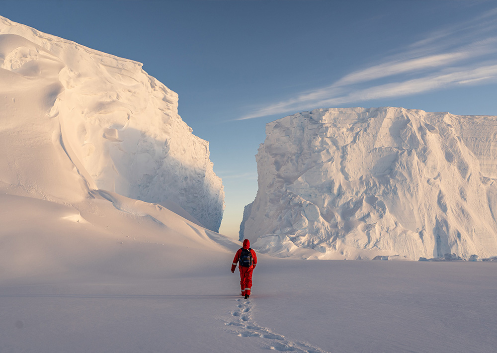 Eine Person in einem roten Anzug, die zur Polarforschung Trautmann gehört, geht durch die Schneelandschaft der Antarktis auf zwei hoch aufragende Eisklippen zu und hinterlässt bei klarem Himmel Fußspuren im Schnee.