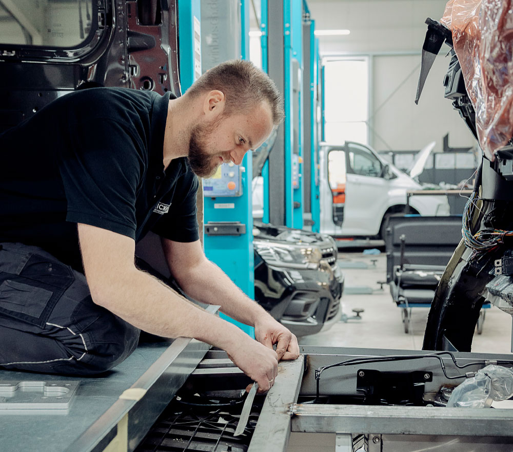 Ein Mann in einem schwarzen Hemd kniet bei Fricke Mobilitätslösungen, einem Spezialisten für Behindertenfahrzeuge, während er an der Innenausstattung eines Fahrzeugs arbeitet. Er benutzt ein Maßband und Werkzeuge, während teilmontierte Fahrzeuge und blaue Hebevorrichtungen die Werkstatt füllen.