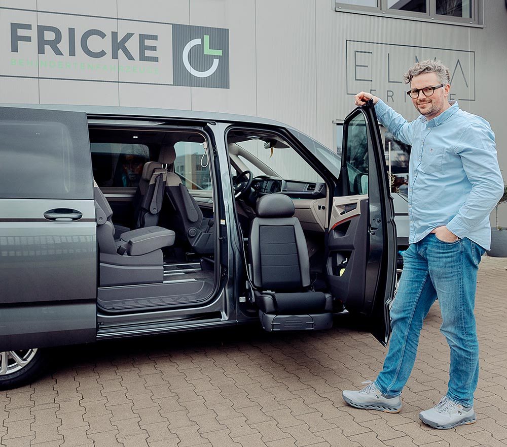 Ein Mann mit Brille, hellblauem Hemd und Jeans steht neben einem grauen Lieferwagen der Firma Fricke Mobilitätslösungen Behindertenfahrzeuge, bei dem die vordere Tür und die seitliche Schiebetür geöffnet sind, so dass ein drehbarer Sitz zum Vorschein kommt. Im Hintergrund ist ein Gebäude mit FRICKE-Schriftzug zu sehen.