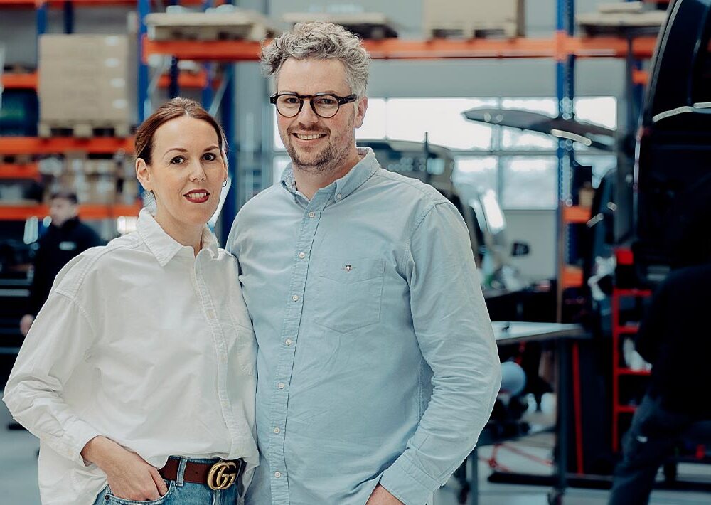 Eine Frau und ein Mann stehen lächelnd in einer Autowerkstatt, die auf Fricke Mobilitätslösungen Behindertenfahrzeuge spezialisiert ist, mit Regalen und Fahrzeugen im Hintergrund und einem Mechaniker, der in der Nähe an einer Werkbank arbeitet.