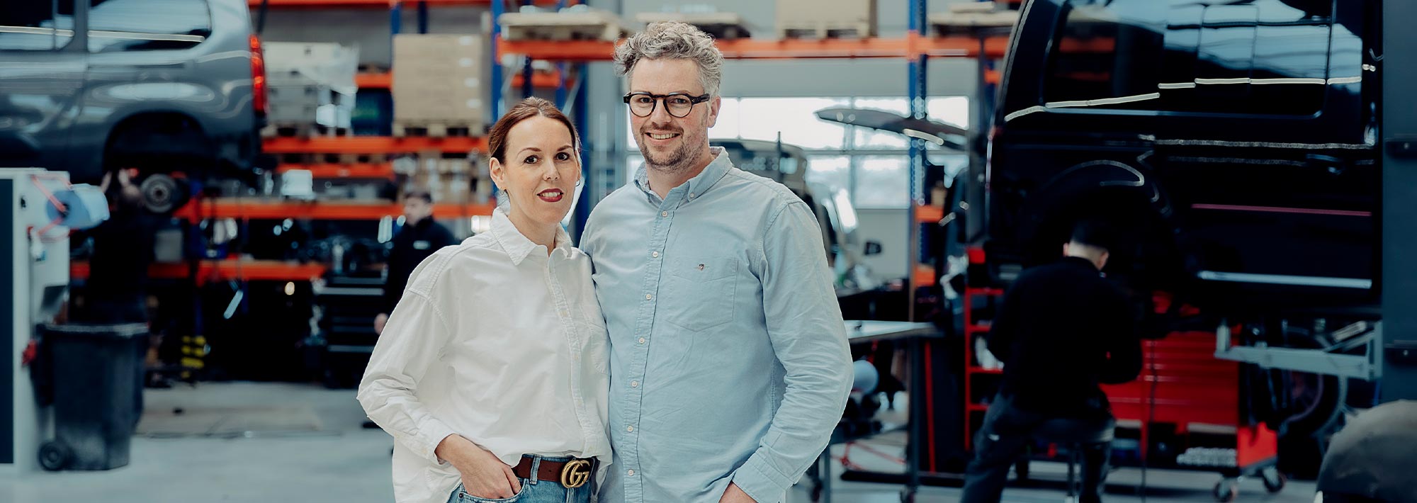 Eine Frau und ein Mann stehen lächelnd in einer Autowerkstatt, die auf Fricke Mobilitätslösungen Behindertenfahrzeuge spezialisiert ist, mit Regalen und Fahrzeugen im Hintergrund und einem Mechaniker, der in der Nähe an einer Werkbank arbeitet.