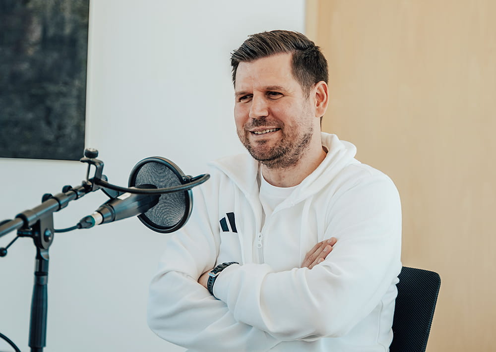 Ein Mann in einem weißen RETERNITY Mode-Kapuzenpulli sitzt mit verschränkten Armen an einem Schreibtisch und spricht lächelnd in ein professionelles Mikrofon während eines Podcasts über Unternehmertum und Gründung.