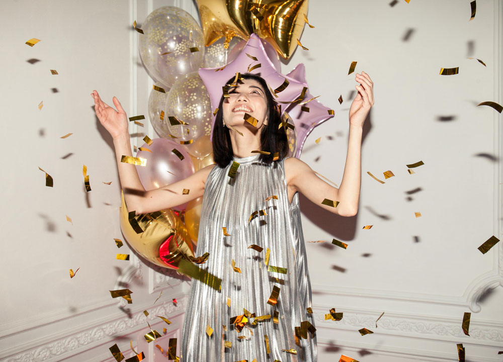 Eine Frau in einem silberglänzenden Kleid lächelt und hebt die Arme, während um sie herum goldenes Konfetti fällt. Hinter ihr sind bunte Luftballons in Gold, Rosa und Transparent zu sehen, die eine festliche Atmosphäre schaffen, die perfekt zu einer Feier des Online-Magazins passt.