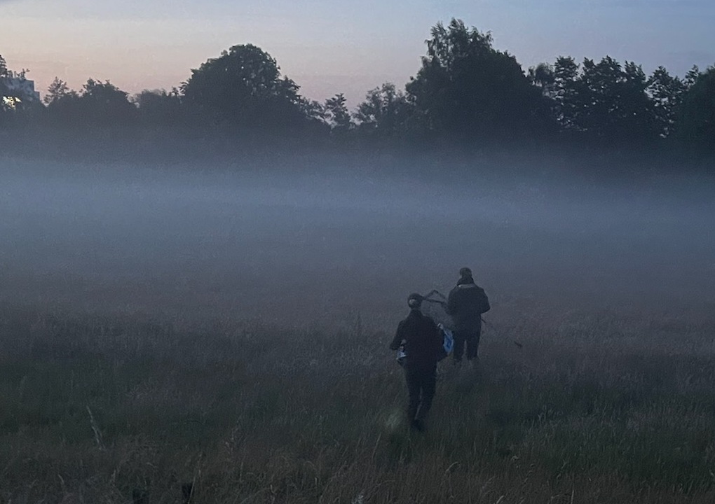 Zwei Menschen gehen in der Dämmerung über eine Wiese, umgeben von Nebel. Im Hintergrund zeichnen sich die Silhouetten von Bäumen vor einem schwach leuchtenden blau-rosa Himmel ab. Eine der beiden Personen trägt ein Fahrrad.