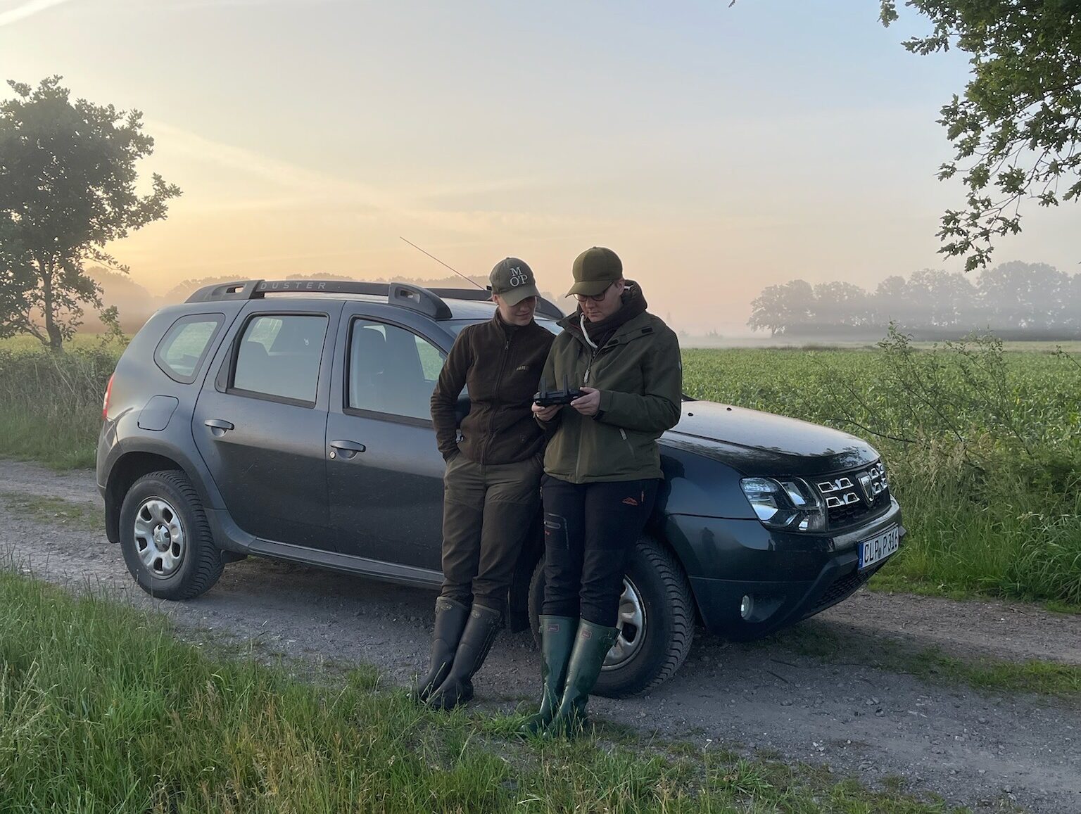 Zwei Personen in Outdoor-Kleidung und Stiefeln stehen neben einem geparkten dunklen Geländewagen auf einer ländlichen Schotterstraße und betrachten gemeinsam ein Gerät. Morgennebel hüllt das Feld und die Bäume im Hintergrund unter einem sanften Sonnenaufgang ein.