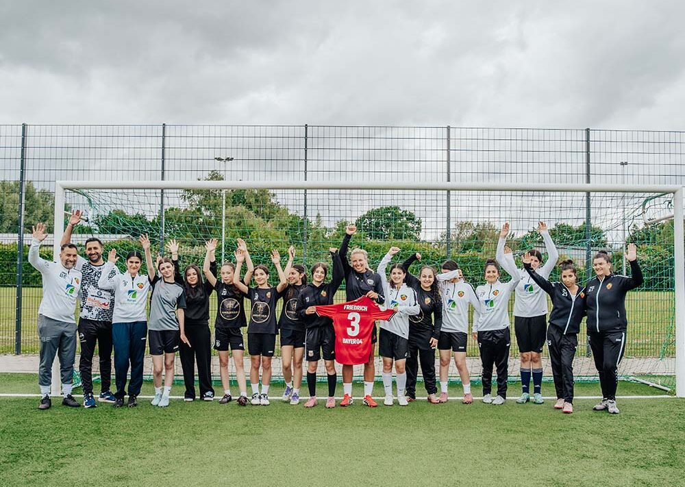 Auf einem Fußballplatz vor einem Tor steht eine Gruppe von Menschen und reckt jubelnd die Hände in die Höhe; eine Person hält dabei ein rotes Trikot mit der Nummer 3 in der Hand.