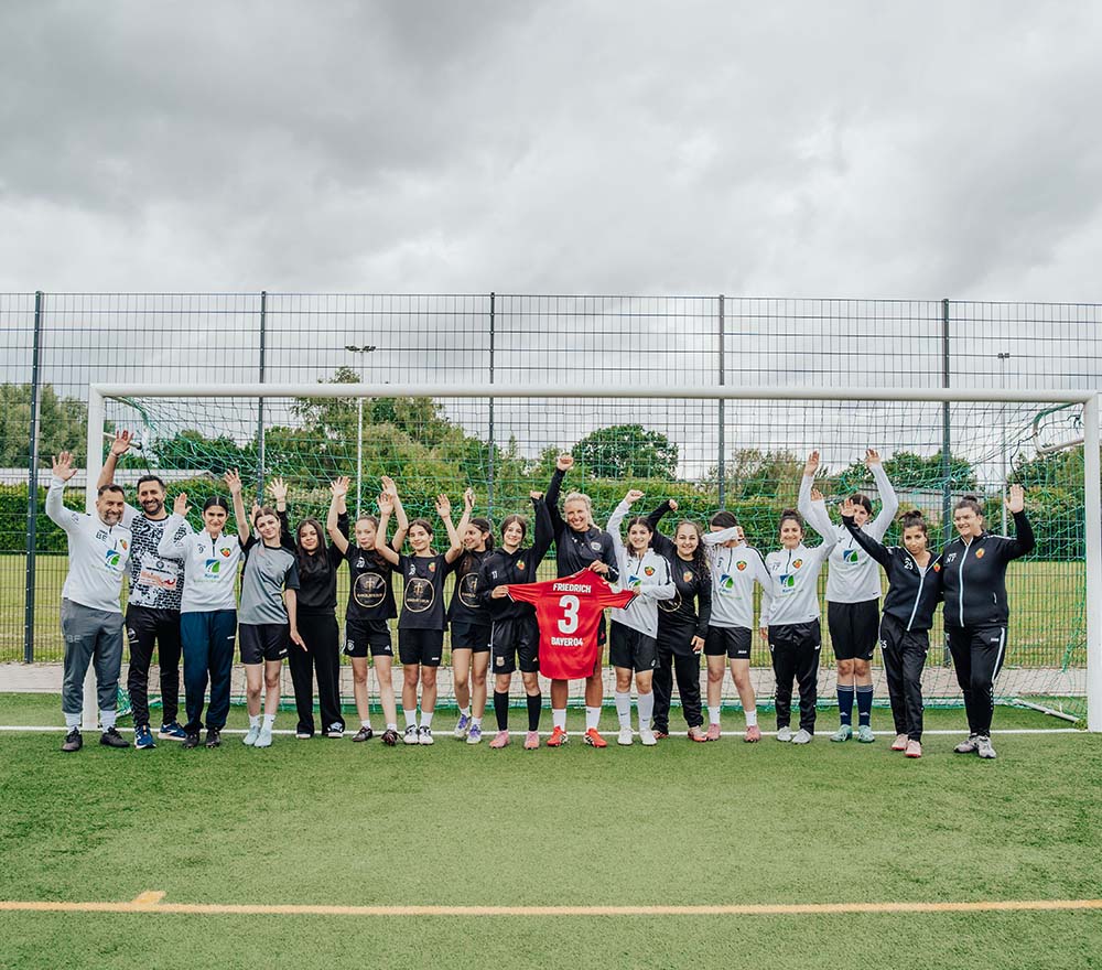 Auf einem Fußballplatz vor einem Tor steht eine Gruppe von Menschen und reckt jubelnd die Hände in die Höhe; eine Person hält dabei ein rotes Trikot mit der Nummer 3 in der Hand.
