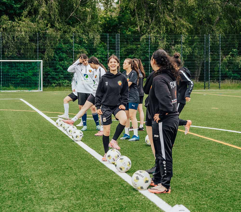 Eine Gruppe von Mädchen in Sportkleidung steht auf einem Fußballplatz, jede mit einem Fußball in der Hand, lächelt und unterhält sich während des Trainings. Im Hintergrund sind Bäume und ein Torpfosten zu sehen.