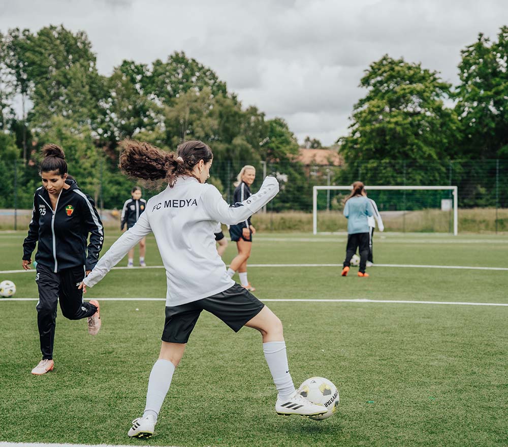 Mädchen spielen Fußball auf einem grünen Feld; ein Mädchen in einem weißen Trikot schießt den Ball, während andere in der Nähe stehen oder herumlaufen. Im Hintergrund sind unter einem bewölkten Himmel Bäume und ein Torpfosten zu sehen.