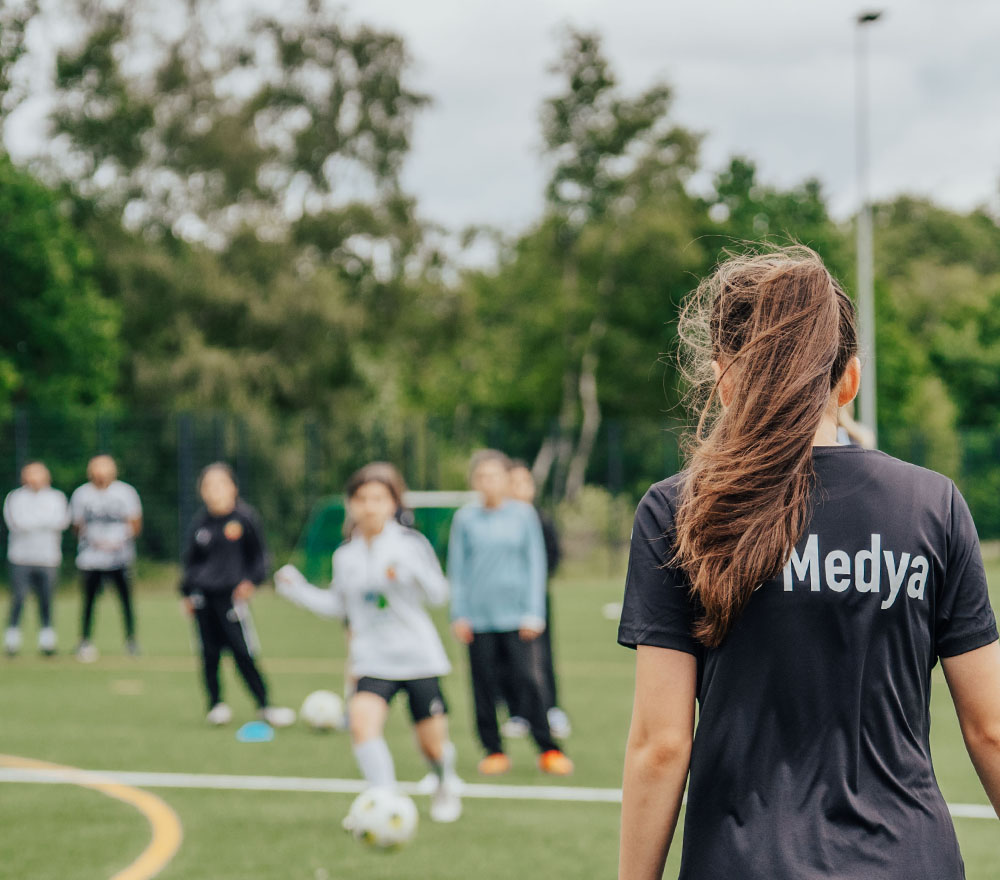 Ein Mädchen mit Pferdeschwanz, das ein Medya-Shirt trägt, schaut sich ein Jugendfußballspiel an, das gerade auf einem grünen Rasen stattfindet; im Hintergrund sind mehrere Kinder und Bäume zu sehen.
