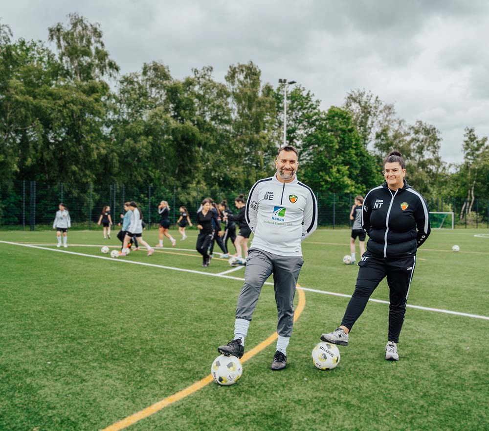 Zwei Fußballtrainer stehen auf einem Spielfeld, wobei jeder mit einem Fuß auf einem Fußball steht, während im Hintergrund eine Gruppe von Spielern trainiert. Das Spielfeld ist von Bäumen umgeben, und der Himmel ist bewölkt.