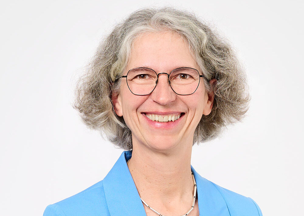 Lächelnde Frau mit grauen Locken, die eine Brille, einen hellblauen Blazer, ein weißes Hemd und eine silberne Halskette trägt und vor einem schlichten weißen Hintergrund posiert – perfekt, um Selbstbewusstsein und Professionalität beim „Ausbildungsstart Finanzen“ zu veranschaulichen.
