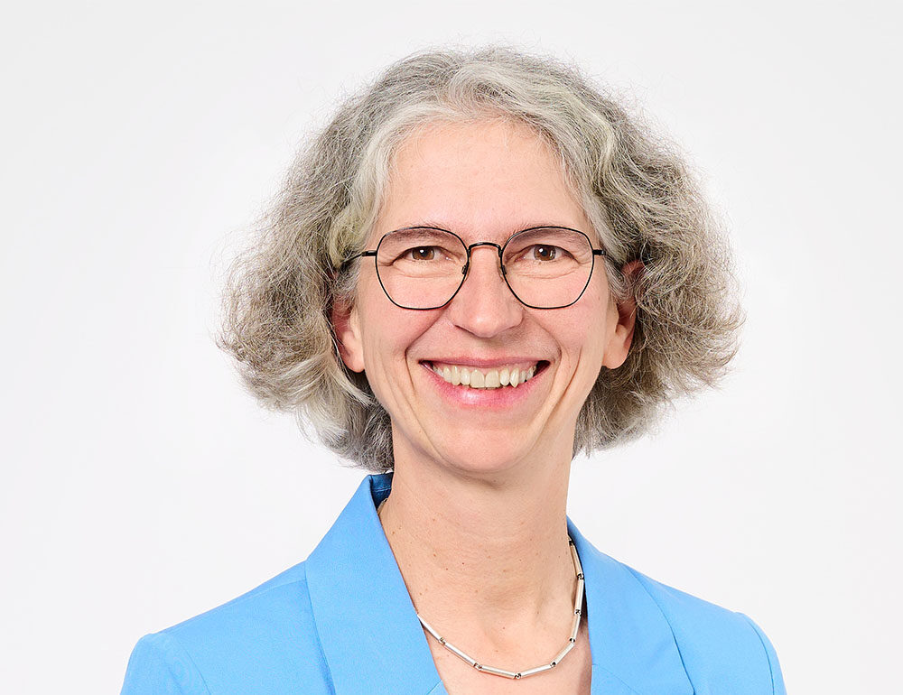 Lächelnde Frau mit grauen Locken, die eine Brille, einen hellblauen Blazer, ein weißes Hemd und eine silberne Halskette trägt und vor einem schlichten weißen Hintergrund posiert – perfekt, um Selbstbewusstsein und Professionalität beim „Ausbildungsstart Finanzen“ zu veranschaulichen.