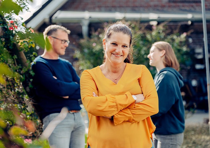 Im Vordergrund steht eine Frau in einer gelben Bluse, die lächelnd die Arme verschränkt hat. Hinter ihr stehen ein Mann und eine weitere Frau im Freien, unterhalten sich und sind leicht unscharf.