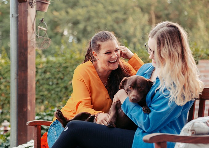 Zwei Frauen sitzen draußen auf einer Bank, lächeln und lachen miteinander. Eine der Frauen hält einen kleinen braunen Hund auf dem Schoß. Im Hintergrund ist viel Grün zu sehen.