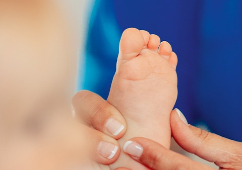 Eine Nahaufnahme eines Erwachsenen, der sanft den kleinen nackten Fuß eines Babys hält. Der Hintergrund ist in Blautönen sanft unscharf.
