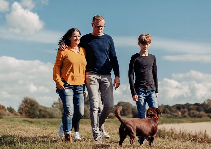 Eine Frau, ein Mann und ein Junge spazieren gemeinsam mit einem braunen Hund draußen auf einer Wiese. Der Himmel ist blau und bewölkt, und im Hintergrund sind Bäume zu sehen.